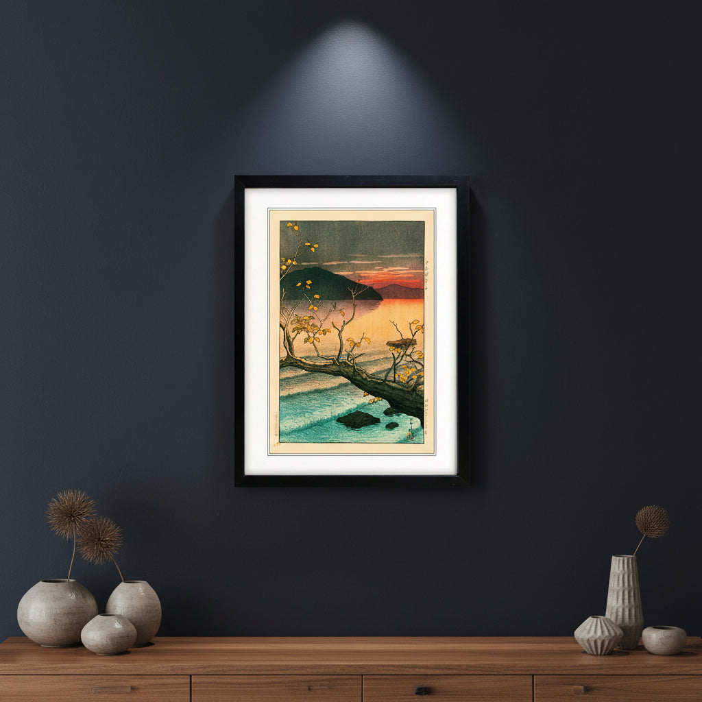 A framed Japanese-style art print depicting a sunset over a calm lake with a dark mountain, a fallen tree branch with yellow leaves, and blue water, shown in a black frame with a white mat on a white background.