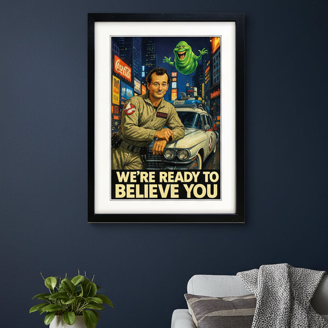A framed retro-style Ghostbusters poster print featuring a man in a beige jumpsuit leaning on a white car, with a green ghost floating above and the text 'WE'RE READY TO BELIEVE YOU' at the bottom. The print is shown in a black frame with a white mat against a light gray background.