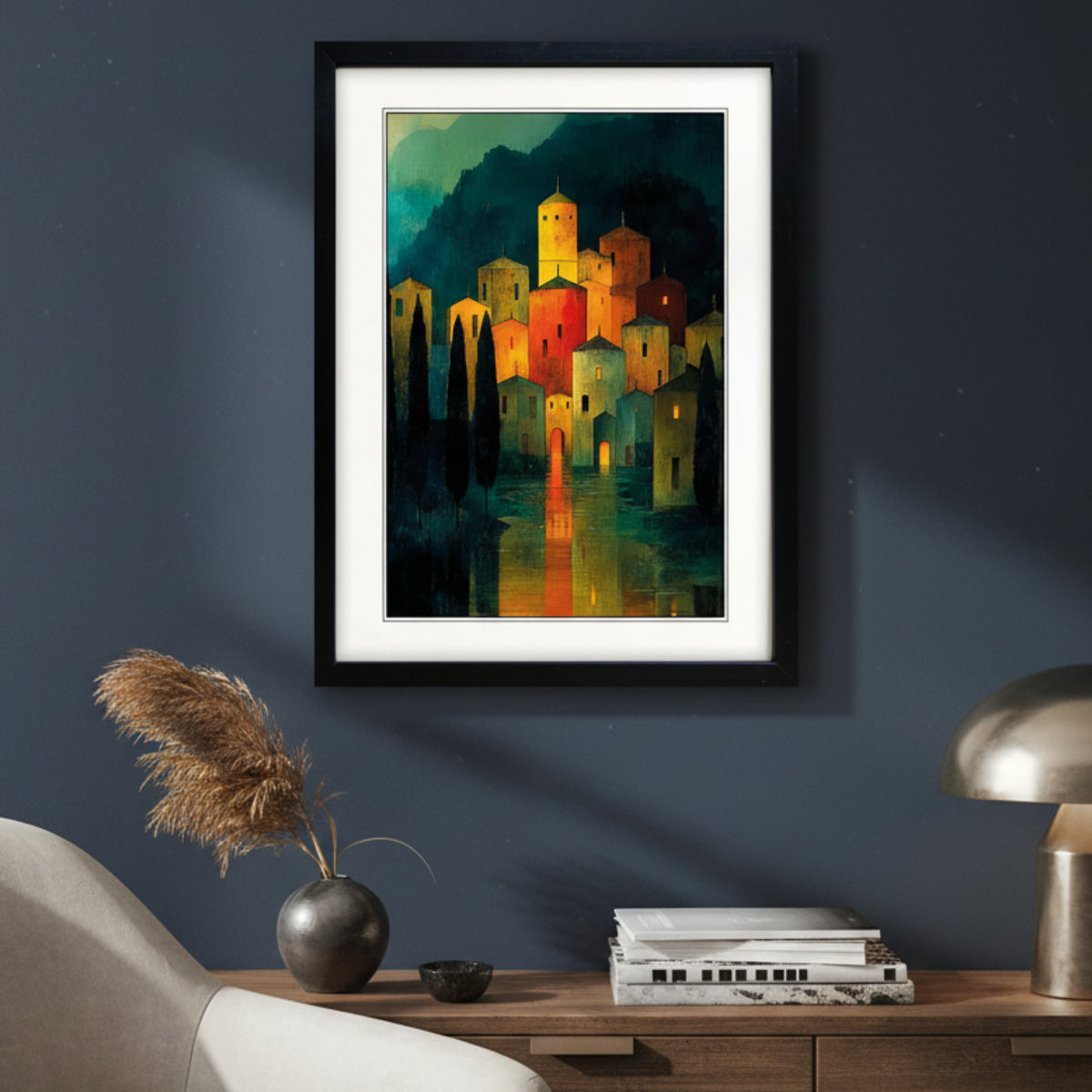 Framed art print featuring a stylized village scene at night with glowing yellow and orange buildings, dark trees, and reflections in water, presented in a black frame with white matting.