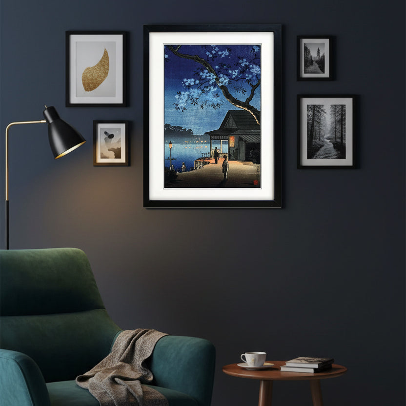 A framed art print featuring a Japanese-style night scene with cherry blossoms, a dock, a small building, and a person standing by the water. The print is matted with a white border and enclosed in a black frame, shown against a plain white background.