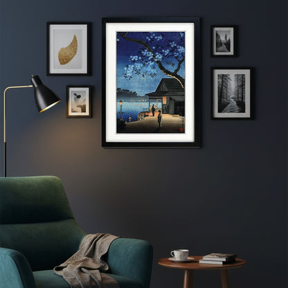 A framed art print featuring a Japanese-style night scene with cherry blossoms, a dock, a small building, and a person standing by the water. The print is matted with a white border and enclosed in a black frame, shown against a plain white background.
