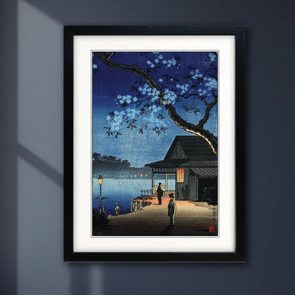 A framed art print featuring a Japanese-style night scene with cherry blossoms, a dock, a small building, and a person standing by the water. The print is matted with a white border and enclosed in a black frame, shown against a plain white background.