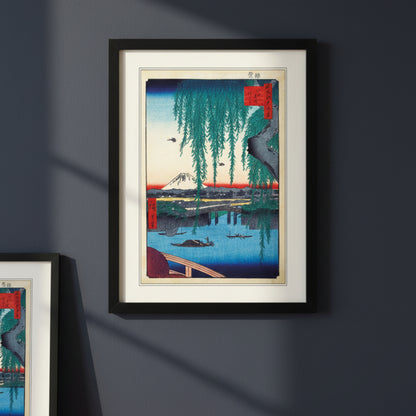 A framed art print featuring a Japanese ukiyo-e woodblock illustration of Mount Fuji, a bridge, boats, and hanging willow branches, presented in a black frame with a white mat on a light gray background.