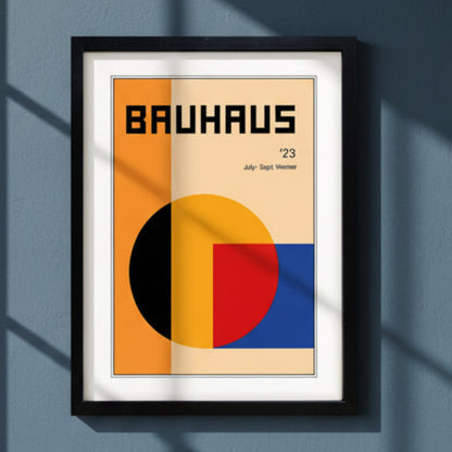 A framed Bauhaus '23 art print featuring a minimalist geometric design with orange, beige, yellow, red, and blue blocks, presented in a black frame against a dark blue wall.