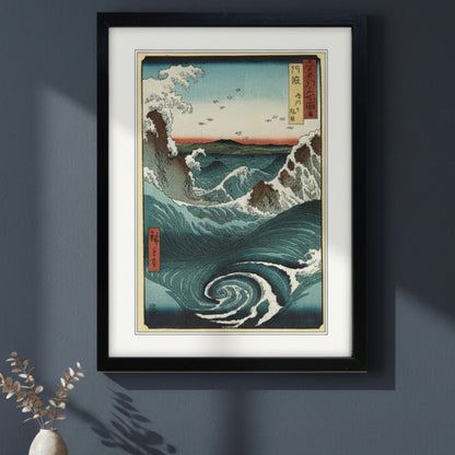 A framed Japanese-style wave art print hangs on a dark blue-gray wall above a wooden console table with a small vase and books. The print shows deep blue ocean waves, white foam, and a pale sky with small birds, enclosed in a black frame with a white mat.