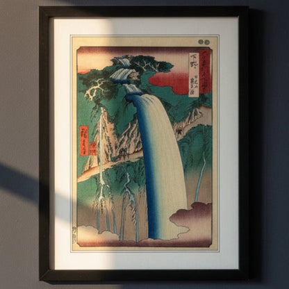 A framed Japanese-style art print depicting a waterfall scene, shown against a plain white background. The artwork features a large cream-colored waterfall, green and blue mountains, red sky accents, and black ink details, enclosed in a black frame with a white mat.