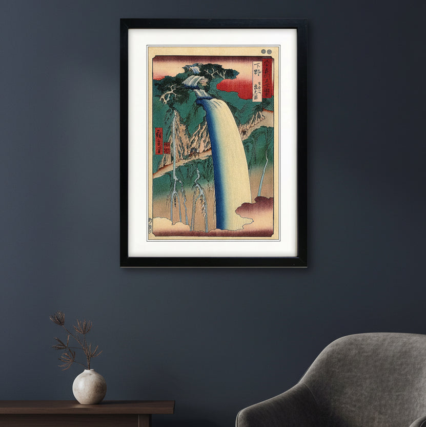 A framed Japanese-style art print depicting a waterfall scene, shown against a plain white background. The artwork features a large cream-colored waterfall, green and blue mountains, red sky accents, and black ink details, enclosed in a black frame with a white mat.