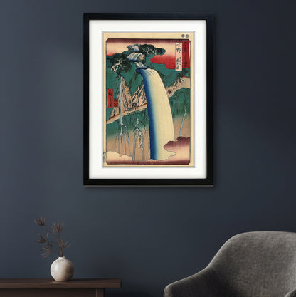 A framed Japanese-style art print depicting a waterfall scene, shown against a plain white background. The artwork features a large cream-colored waterfall, green and blue mountains, red sky accents, and black ink details, enclosed in a black frame with a white mat.