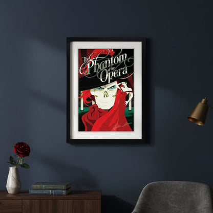 A framed art print of 'The Phantom of the Opera' featuring a stylized illustration of the Phantom skull in a black suit and red cape, with a woman in a red dress in the background, presented in a black frame with a grey wall background