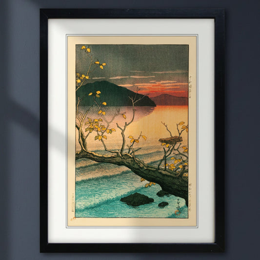 A framed Japanese-style art print depicting a sunset over a calm lake with a dark mountain, a fallen tree branch with yellow leaves, and blue water, shown in a black frame with a white mat on a white background.