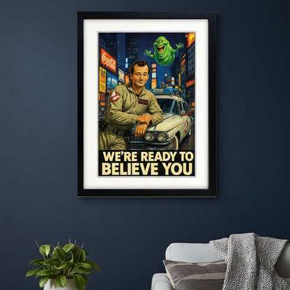 A framed retro-style Ghostbusters poster print featuring a man in a beige jumpsuit leaning on a white car, with a green ghost floating above and the text 'WE'RE READY TO BELIEVE YOU' at the bottom. The print is shown in a black frame with a white mat against a light gray background.