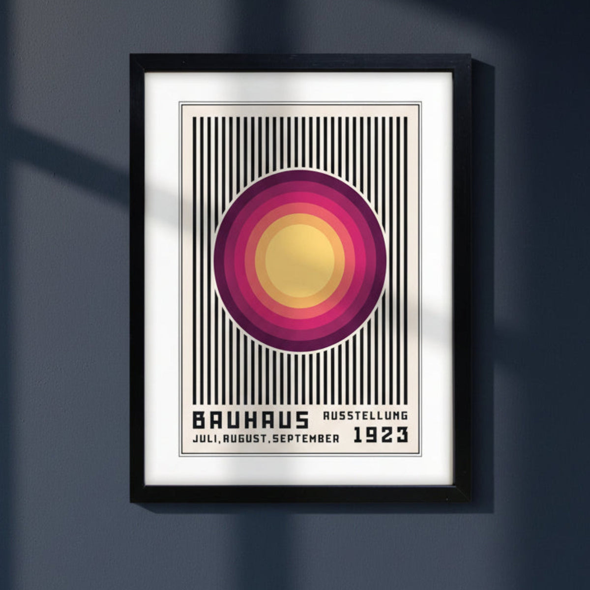 A framed Bauhaus-style art print hanging on a dark gray wall, featuring a central circular motif in yellow, pink, and purple over black vertical stripes, with the text 'BAUHAUS AUSSTELLUNG JULI, AUGUST, SEPTEMBER 1923' at the bottom. The black framed Print is on a dark blue background or wall with natural light.