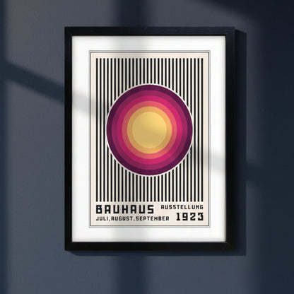 A framed Bauhaus-style art print hanging on a dark gray wall, featuring a central circular motif in yellow, pink, and purple over black vertical stripes, with the text 'BAUHAUS AUSSTELLUNG JULI, AUGUST, SEPTEMBER 1923' at the bottom. The black framed Print is on a dark blue background or wall with natural light.