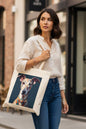 A woman wearing a cream linen shirt and blue jeans carries a canvas tote bag with a custom printed portrait of a greyhound dog on a dark background, set against a blurred city street backdrop.