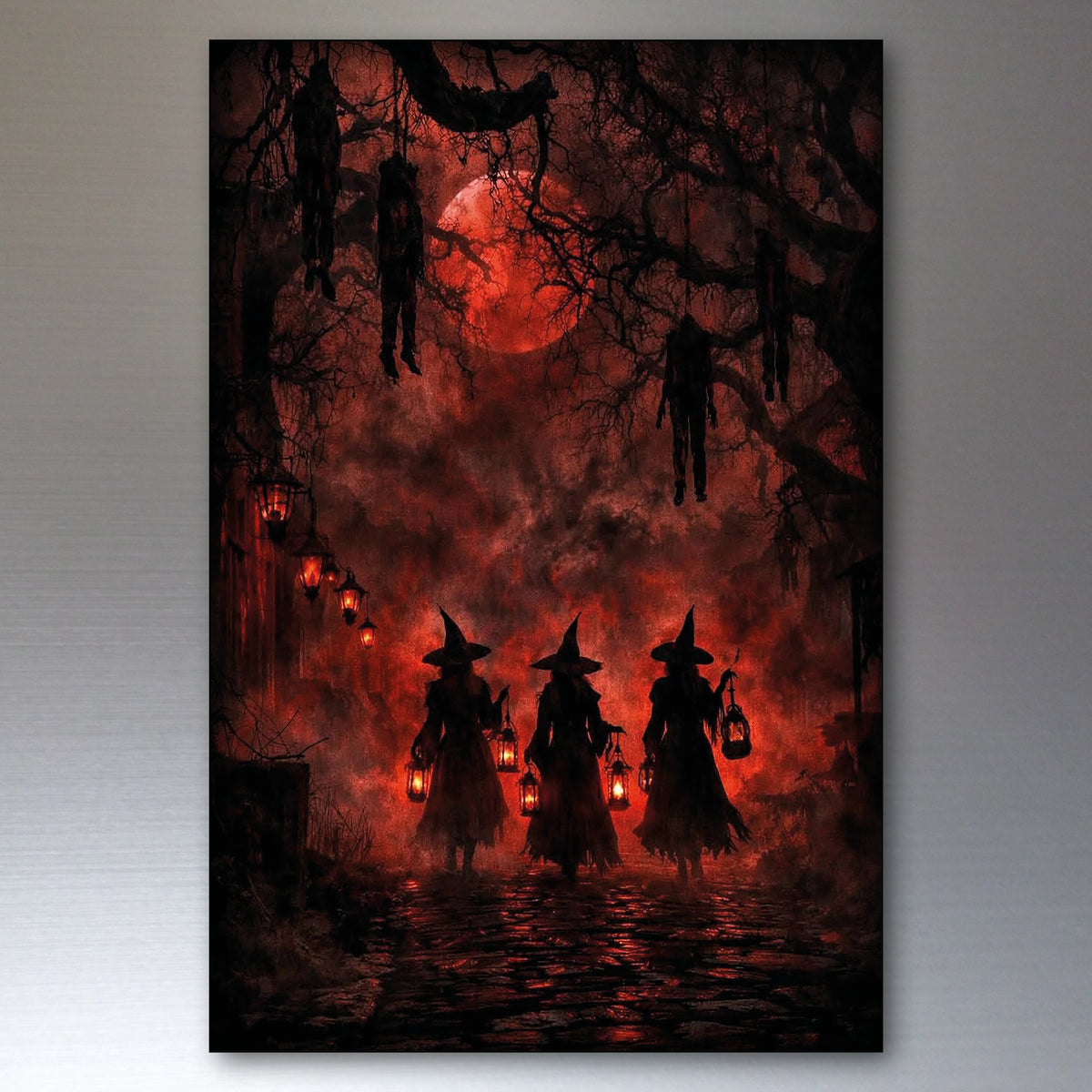 A vertical rectangular wall art print or canvas showing a dark Halloween scene: three witch silhouettes holding lanterns under bare trees with hanging decorations, a glowing red full moon, and red misty sky. The artwork is photographed against a light gray wall.