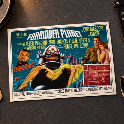 A vintage-style movie poster for 'Forbidden Planet' mounted on a brushed metal wall. The poster features a teal and blue space-themed design with illustrated and photographed figures, including a large robot and a woman in a yellow dress, plus M-G-M branding and film credits.