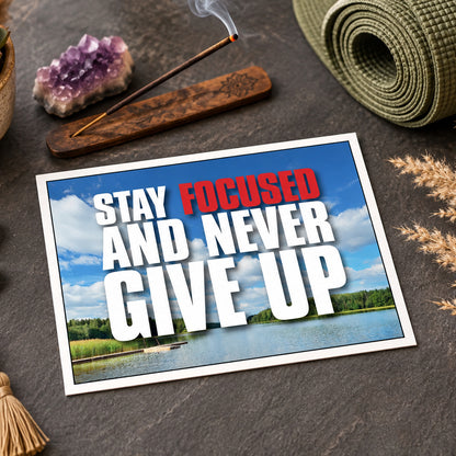 A printed motivational card or poster with the text 'STAY FOCUSED AND NEVER GIVE UP' over a lake landscape, styled on a dark stone surface with an incense stick, crystal, rolled mat, and dried grass.