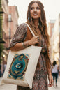 A cream-colored canvas tote bag featuring a large, detailed graphic print of an all-seeing eye design in blue, gold, and red on the front, young bohemian hip women wearing bag showing bag to the side lifestyle