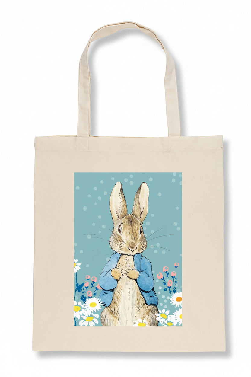 A natural canvas tote bag featuring a printed illustration of Peter Rabbit in a blue jacket surrounded by white daisies on a light blue background.
