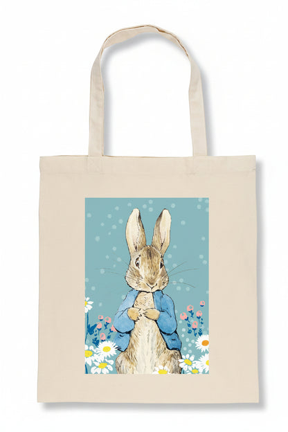 A natural canvas tote bag featuring a printed illustration of Peter Rabbit in a blue jacket surrounded by white daisies on a light blue background.