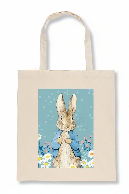 A natural canvas tote bag featuring a printed illustration of Peter Rabbit in a blue jacket surrounded by white daisies on a light blue background.