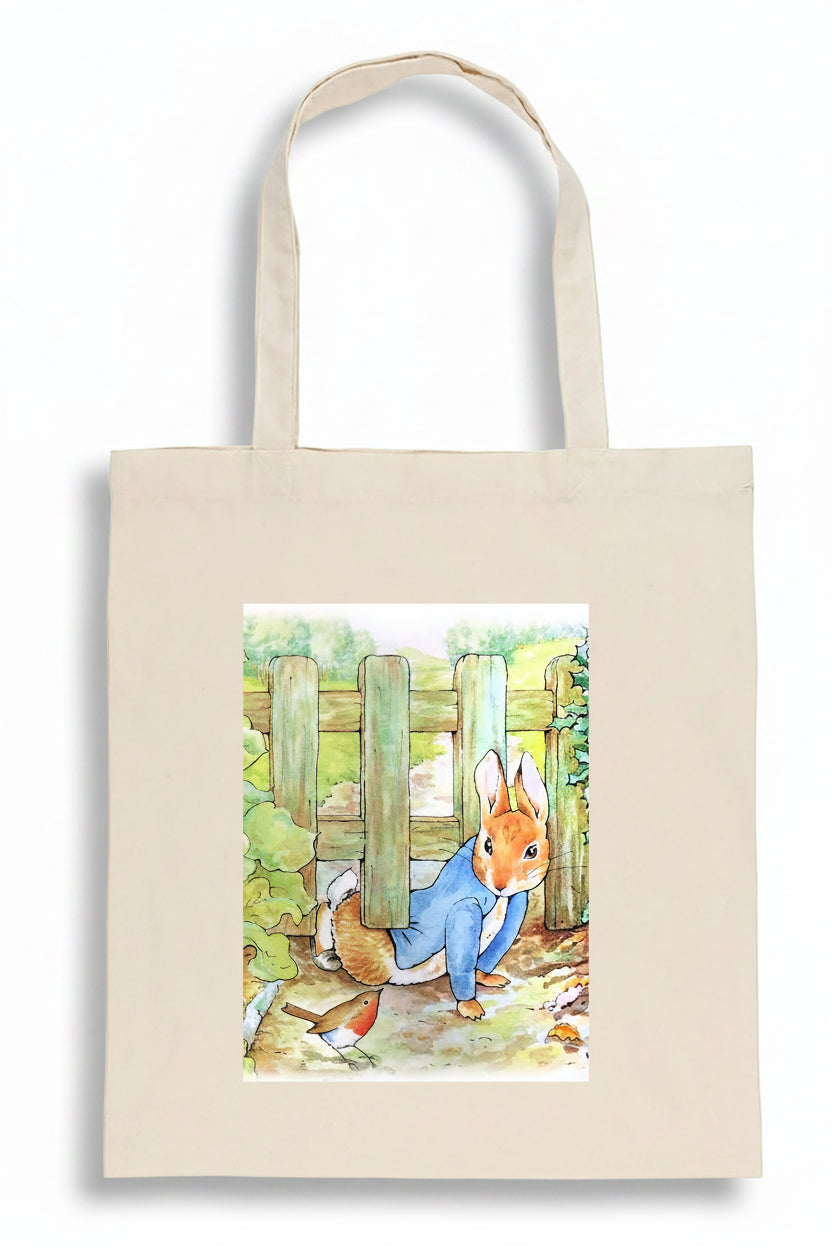 A natural canvas tote bag featuring a watercolor illustration of Peter Rabbit sitting in front of a wooden fence, set against a plain white background.
