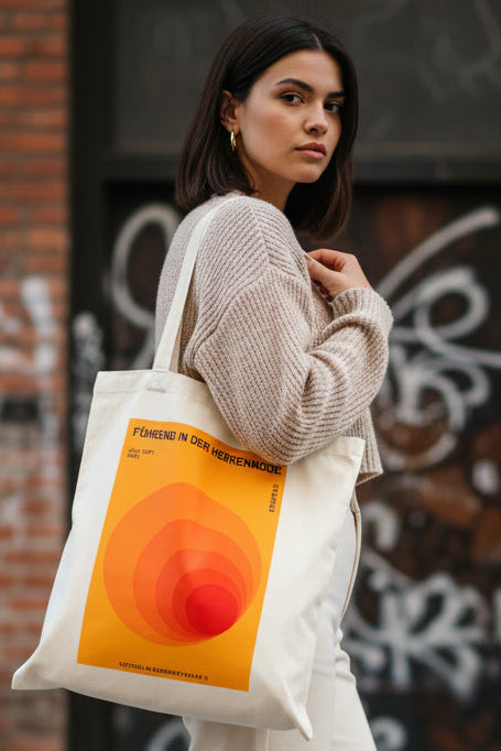 A cream-colored canvas tote bag featuring a retro-style orange and yellow graphic print with German text, displayed against a solid white background.
