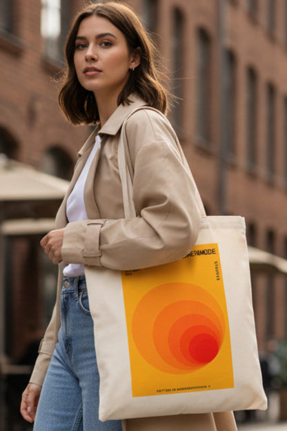 A cream-colored canvas tote bag featuring a retro-style orange and yellow graphic print with German text, displayed against a solid white background.