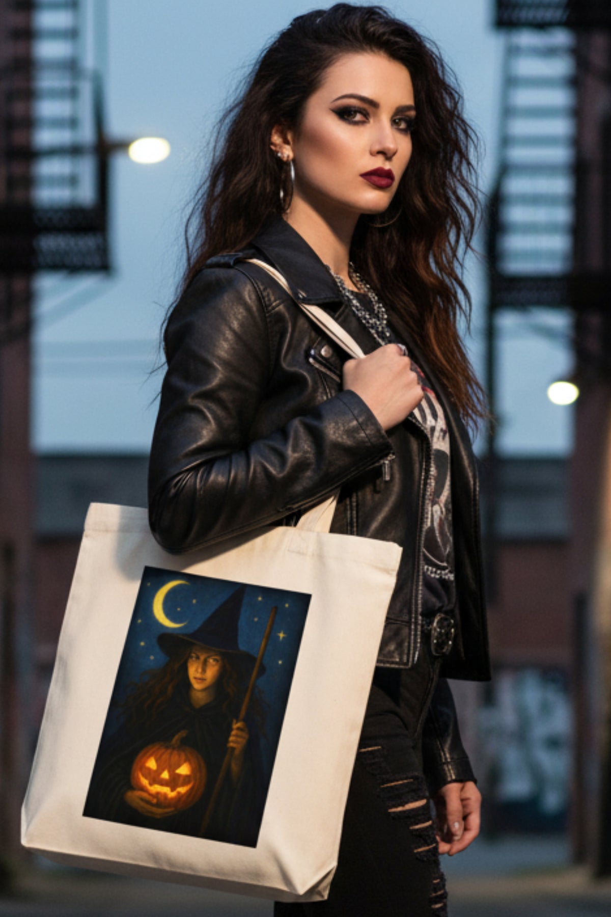 An off-white canvas tote bag featuring a rectangular graphic print of a witch holding a glowing jack-o'-lantern under a crescent moon and stars.