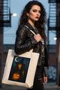 An off-white canvas tote bag featuring a rectangular graphic print of a witch holding a glowing jack-o'-lantern under a crescent moon and stars.