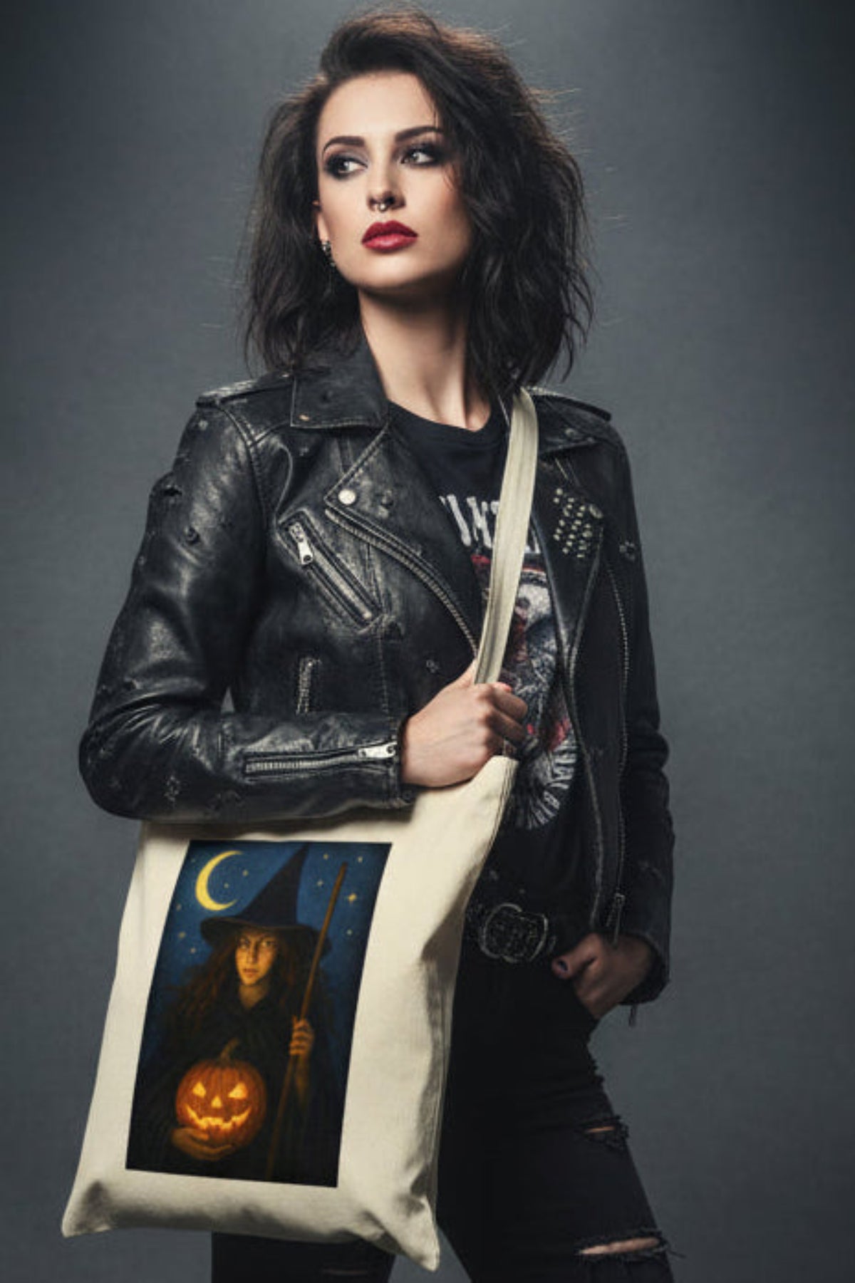 An off-white canvas tote bag featuring a rectangular graphic print of a witch holding a glowing jack-o'-lantern under a crescent moon and stars.